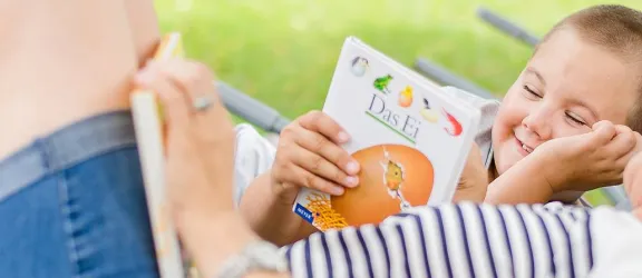 Betreuerin liegt neben Kind in Hängematte und zeigt ein Buch
