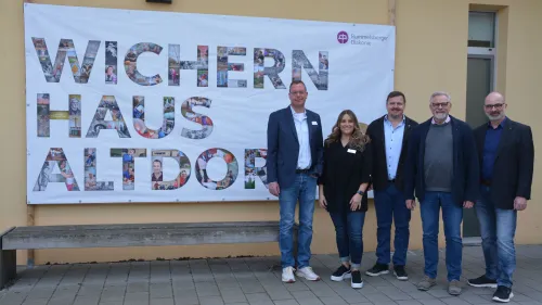 Thorsten Dierolf, Marisa Opel, Martin Tabor, Diakon Volker Deeg und Diakon Thomas Jacoby vor dem neuen Plakat an den Wichernhaus Werkstätten. Anlass ist das 100. Jubiläum der Einrichtung.